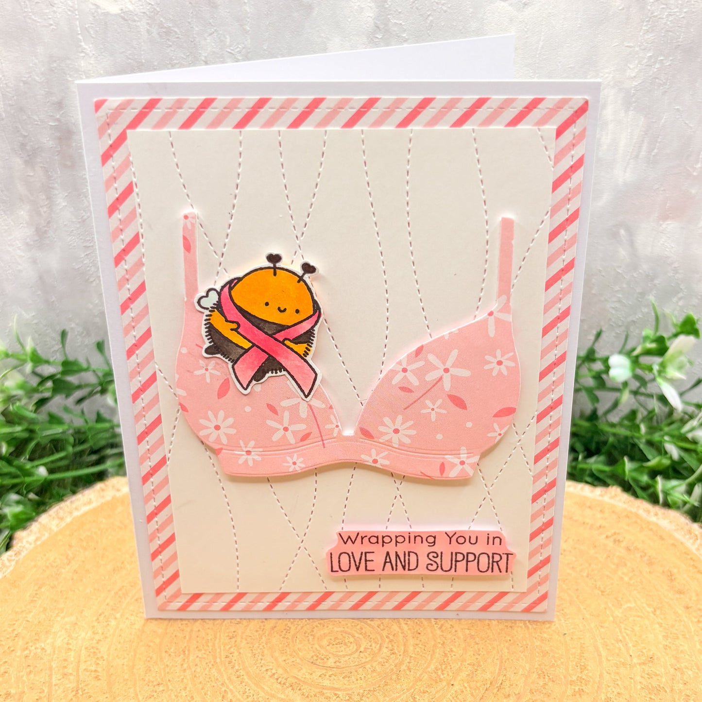 Card with a bra design and bee holding a pink ribbon, on a textured surface with plants in the background. Breast Cancer Awareness Bee & Bra Handmade Support Card