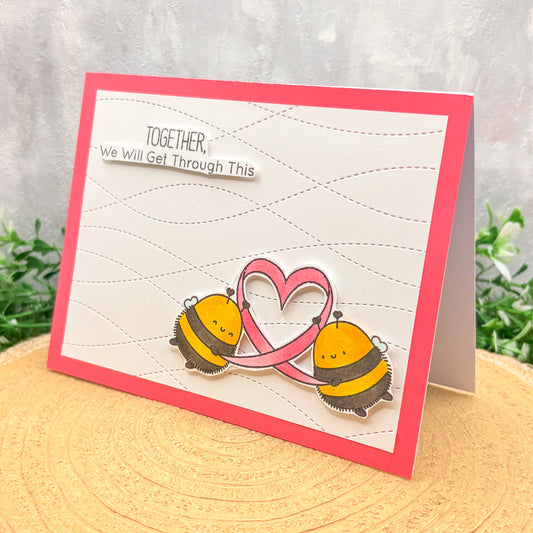 Card with two bees holding a heart and text 'Together, we will get through this' on a textured surface.