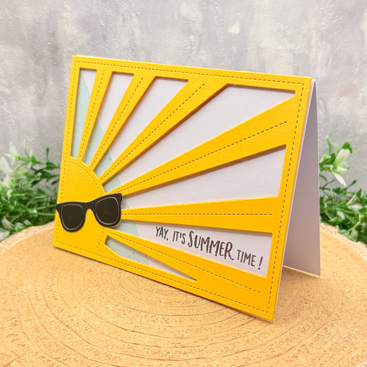 Yellow sun-shaped card with sunglasses and 'Yay, it's Summer time!' text on a wooden surface with plants in the background.