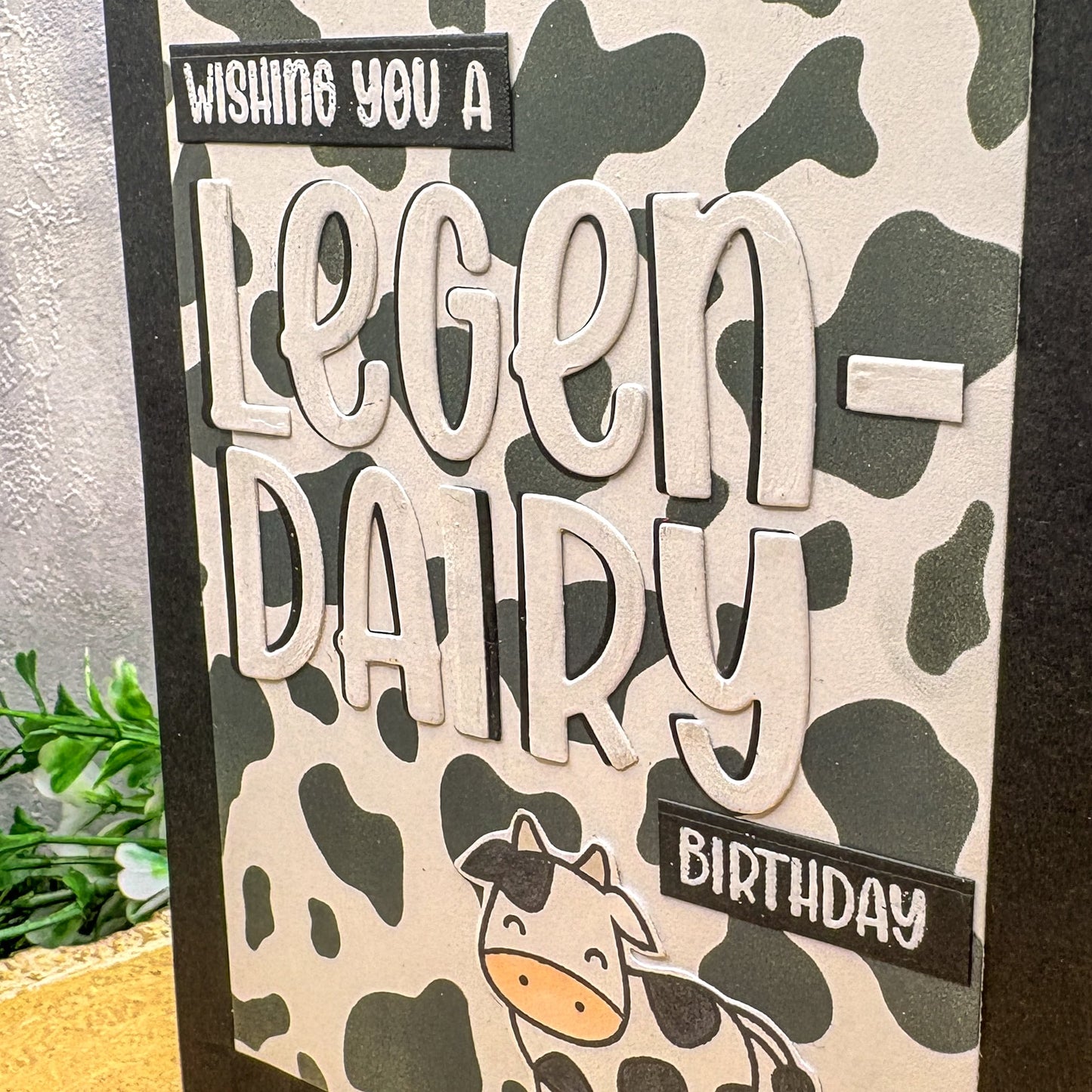 Birthday card with cow pattern and text 'Wishing you a legen-dairy birthday' on a camouflage background.