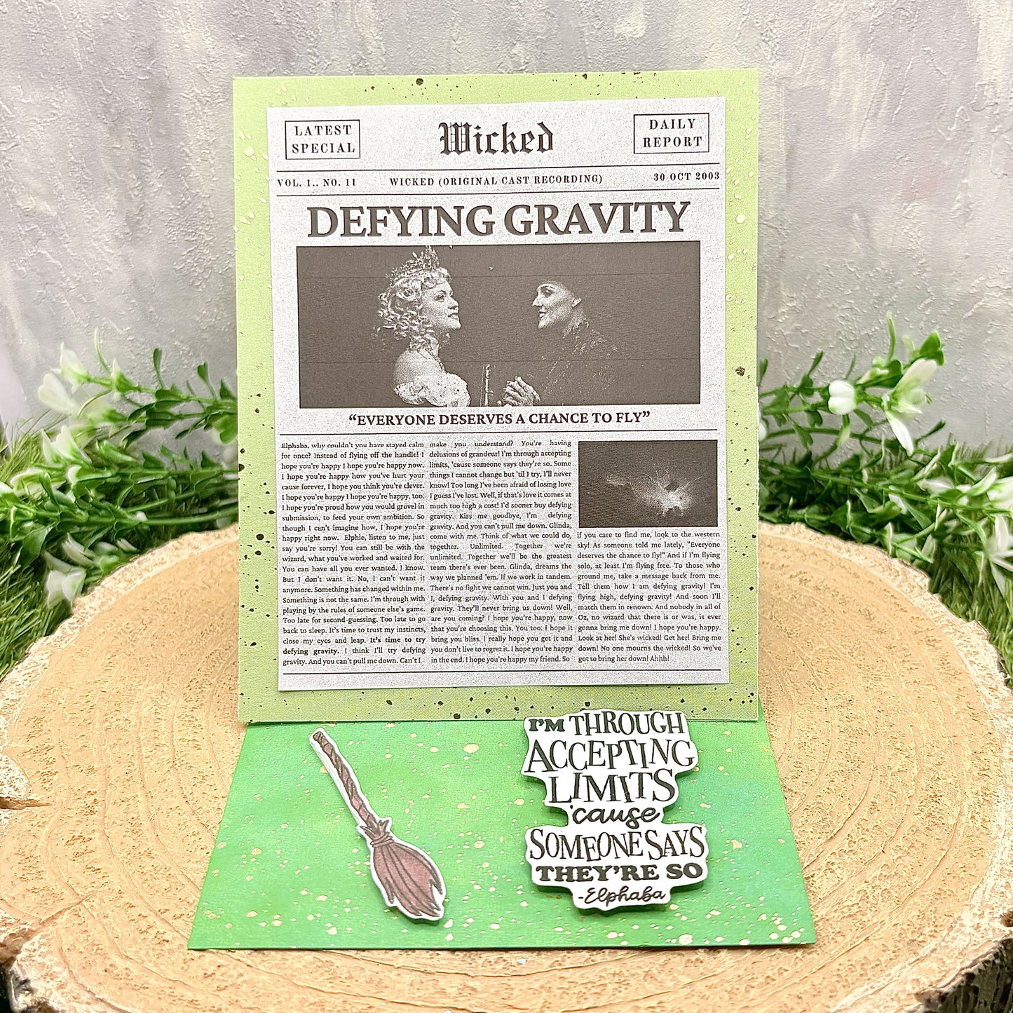 Newspaper-style card with 'Wicked' theme and green border on a wooden surface with plants.