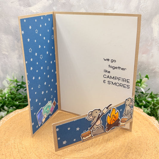 Card with cartoon characters by a campfire and text 'we go together like CAMPFIRE & S'MORES' on a blue and white background.