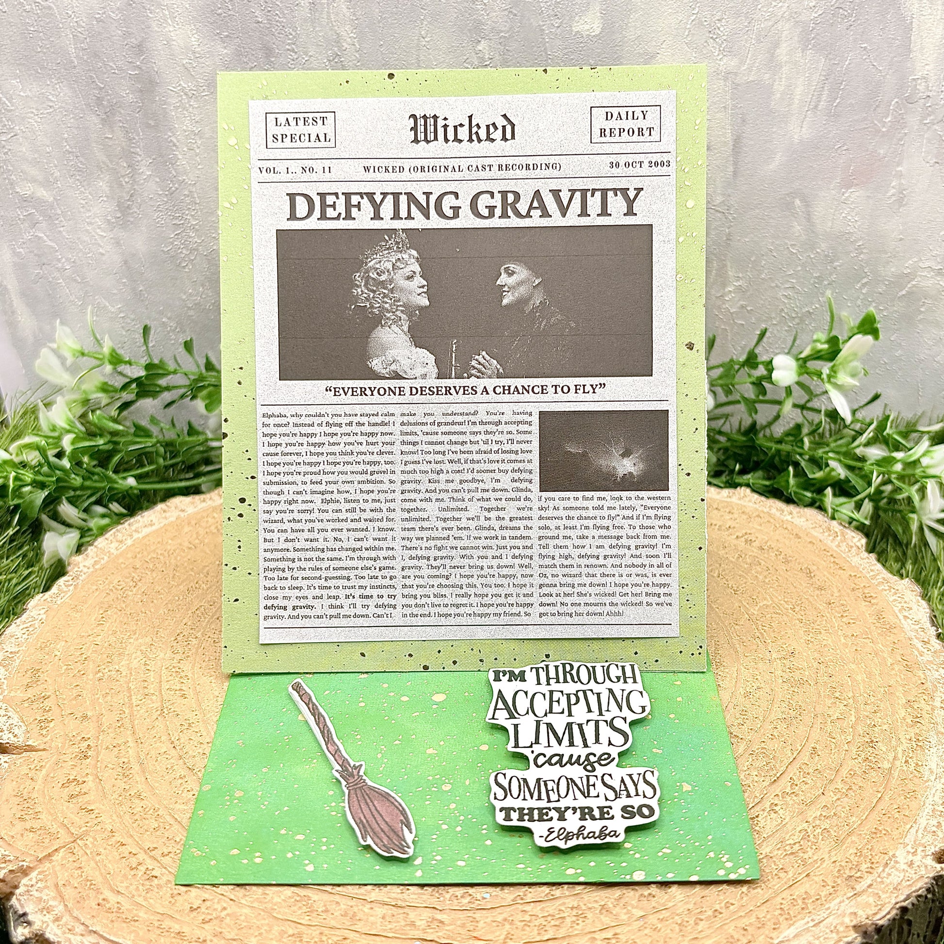 Newspaper-style card with 'Wicked' theme and green border on a wooden surface with plants.