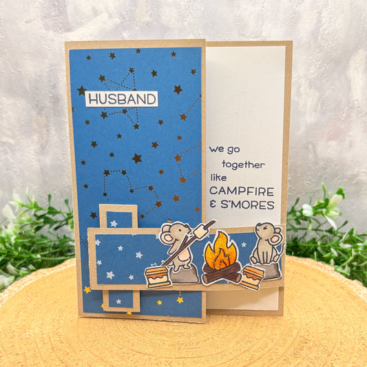 Greeting card for a husband with cartoon bears around a campfire and text 'we go together like CAMPFIRE & S'MORES' on a blue background.